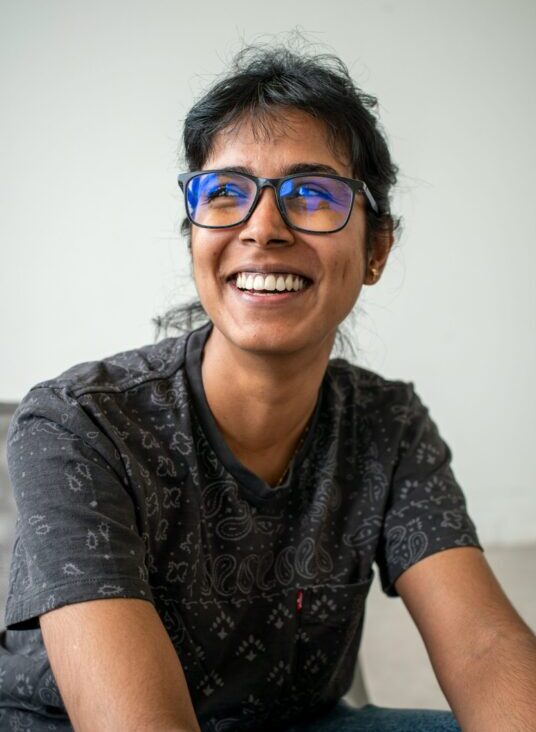 Dark haired woman in dark glasses and a grey shirt smiling while looking off to the side
